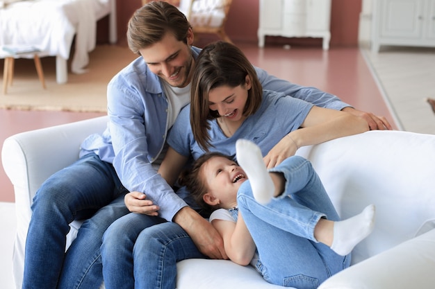 Padres jóvenes amables positivos con una pequeña hija sonriente sentada en el sofá y divirtiéndose juntos en casa.