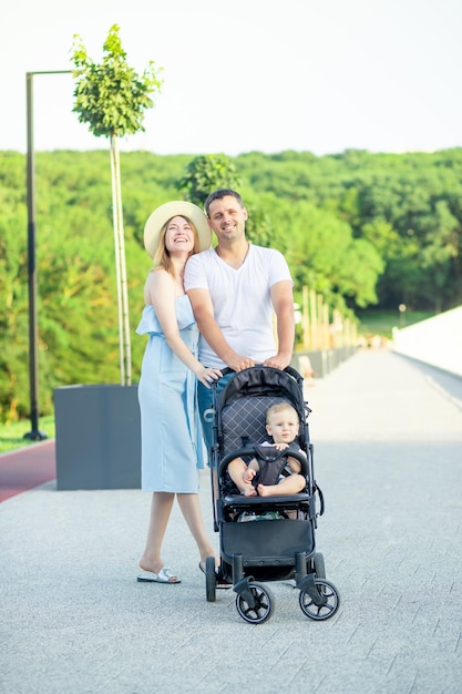 Los padres jóvenes felices mamá y papá están caminando con un bebé en un cochecito en el parque con un sombrero en el verano en la puesta de sol y sonriendo divirtiéndose