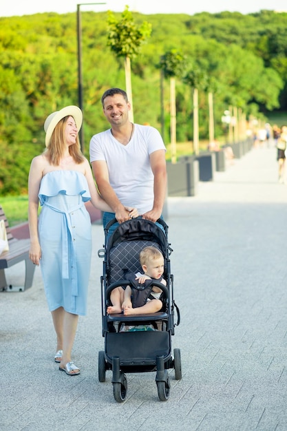 Los padres jóvenes felices mamá y papá están caminando con un bebé en un cochecito en el parque con un sombrero en el verano en la puesta de sol y sonriendo divirtiéndose