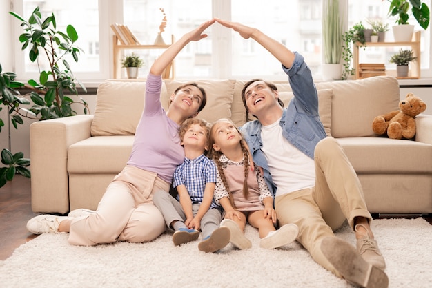 Los padres jóvenes manteniendo sus manos cerca formando un techo sobre sus dos hijos pequeños mientras están sentados en el piso de la sala de estar