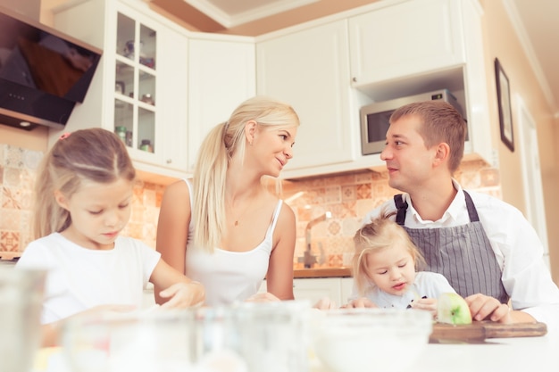 Los padres jóvenes y sus dos niñas hermosas y lindas niñas cocinando y desayunando