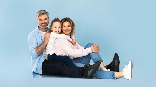 Padres de mediana edad e hija abrazándose sentados sobre fondo azul.