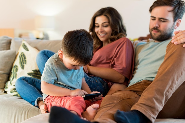 Los padres miran a su hijo usando el teléfono inteligente mientras están sentados en el sofá en casa