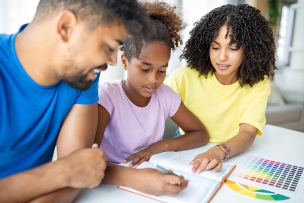 Padres multiétnicos ayudando a su hija con su tarea en casa Padre y madre jóvenes ayudando a su hija a estudiar en la sala de estar Niña completando sus ejercicios con la ayuda de papá y mamá