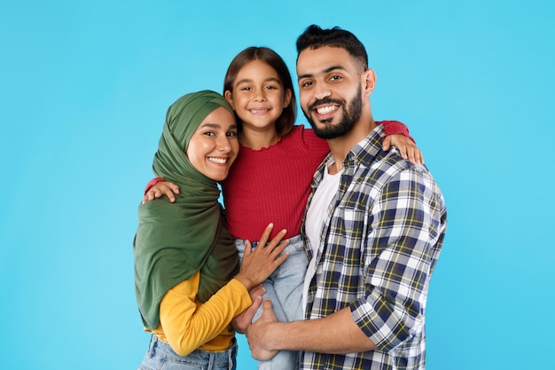 Los padres musulmanes abrazando a la pequeña hija posando juntos sobre fondo azul.