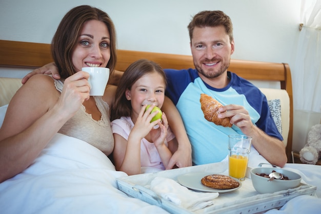 Padres sentados en la cama con su hija y desayunando