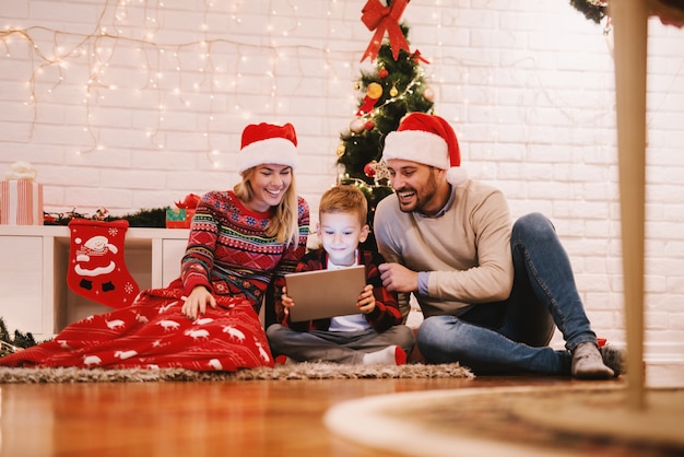 Los padres y su hijo sentados en el piso frente al árbol de Navidad. Niño con tableta y ver videos. Concepto de vacaciones de navidad.