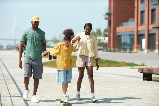 Padres tomándose de la mano con su hijo y enseñándole a andar en patineta en el parque