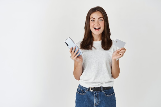 Pagamento com cartão de comércio eletrônico jovem feliz segurando cartão de crédito plástico e smartphone olhando para a câmera surpresa em pé sobre fundo branco