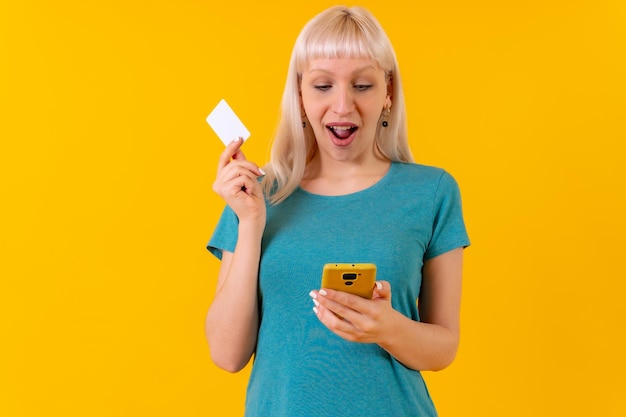 Pagamento on-line com o cartão no telefone menina caucasiana loira no estúdio em um fundo amarelo