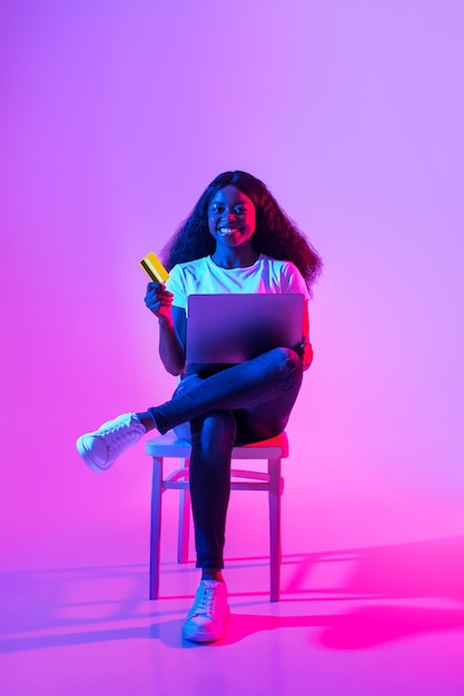 Pagamentos online feliz mulher afro-americana com cartão de crédito e laptop sentado na cadeira em neon