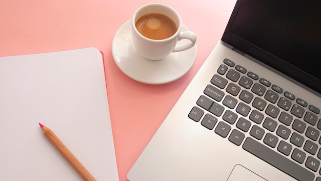 Página en blanco de un cuaderno junto a un portátil y una taza blanca con café sobre un fondo rosa claro