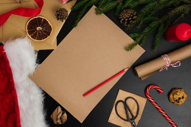 Página de papel castanho em branco em madeira preta Tábua com decoração de Natal Mock Up de carta para Papai Noel Vista superior de Jolly Father Mesa com envelopes Ramos de abeto Ornamento de Natal Vista de cima