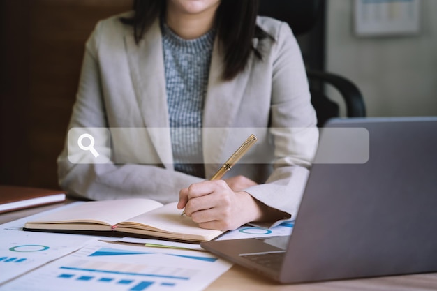 Página de pesquisa feminina trabalhando à mão no computador e escrevendo em um bloco de notas com uma caneta no escritório na mesa de madeira, há um diagrama de negócios gráfico