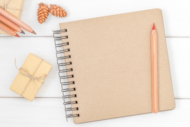 Página vacía del cuaderno para escribir con lápices, caja de regalo y conos sobre fondo blanco de madera