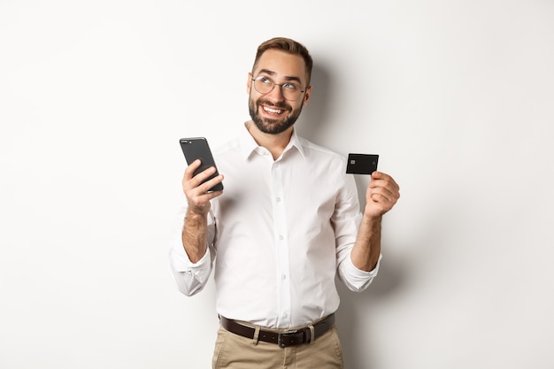 Pago comercial y online. Imagen de hombre guapo pensando mientras sostiene la tarjeta de crédito y el teléfono inteligente, de pie contra el fondo blanco.