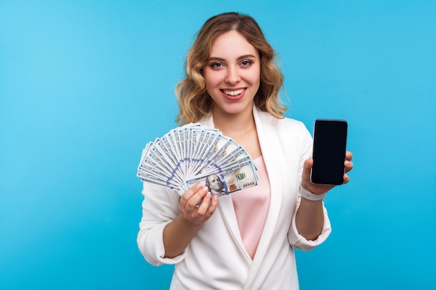 Pago en línea, transacción de dinero. Retrato de mujer positiva con cabello ondulado en chaqueta blanca que muestra dólares y teléfono móvil, con ganancias de internet,. tiro del estudio aislado sobre fondo azul