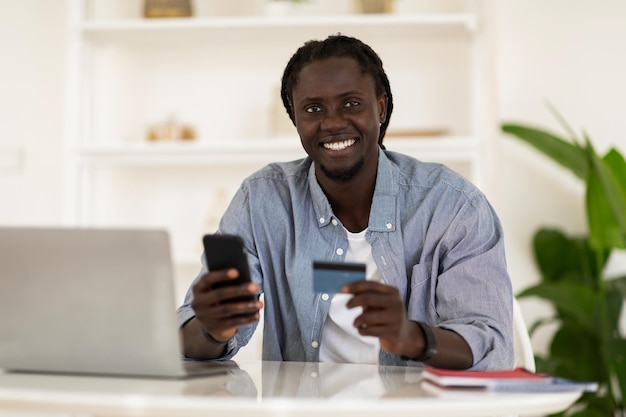 Pagos en línea Sonriente hombre negro independiente usando teléfono inteligente y tarjeta de crédito