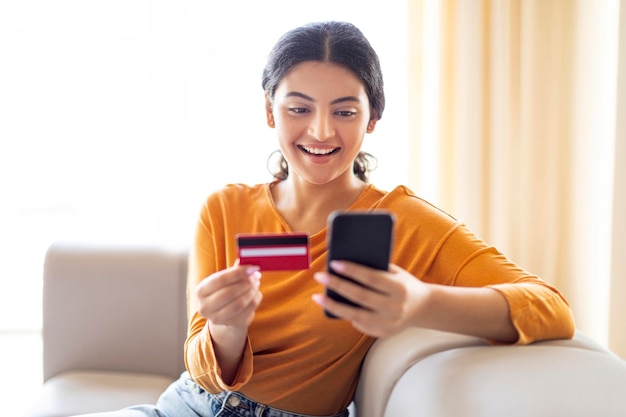 Pagos móviles hermosa mujer india usando teléfono inteligente y tarjeta de crédito en casa