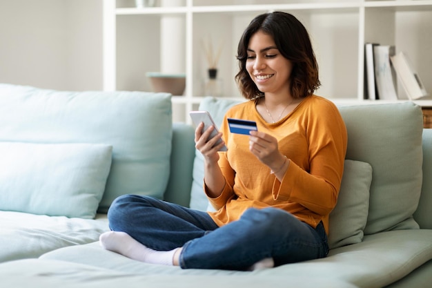 Pagos móviles Mujer árabe joven usando teléfono inteligente y tarjeta de crédito en casa