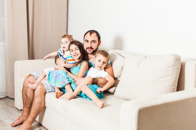 Pai abraça seus três filhos. Retrato de jovem com crianças sentadas no sofá.