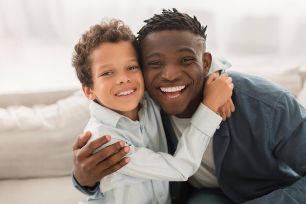 pai afro-americano abraçando filho pequeno olhando para a câmera dentro de casa