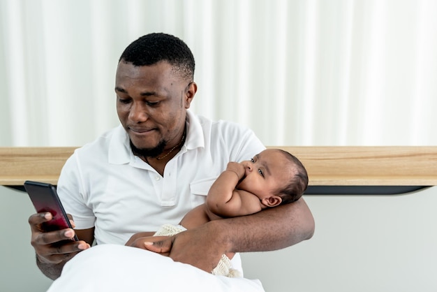 Pai afro-americano atualmente usando o telefone celular enquanto segura um bebê de 3 meses recém-nascido