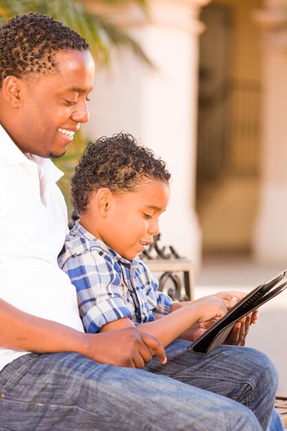 Pai afro-americano e filho mestiço usando tablet de computador no banco do parque