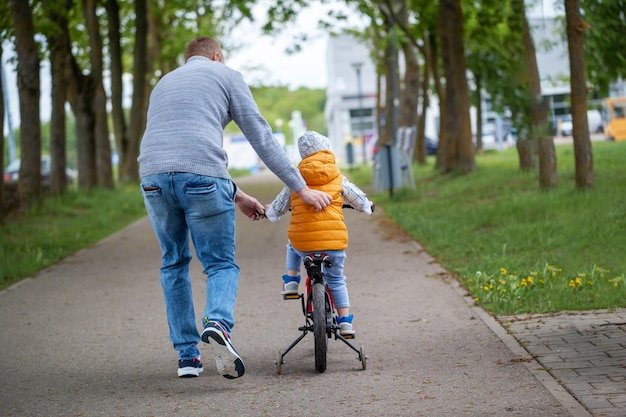 Pai ajuda filho a andar de bicicleta no parque de verão. Garoto andando de bicicleta no parque ao lado de seu pai por trás. Vista traseira