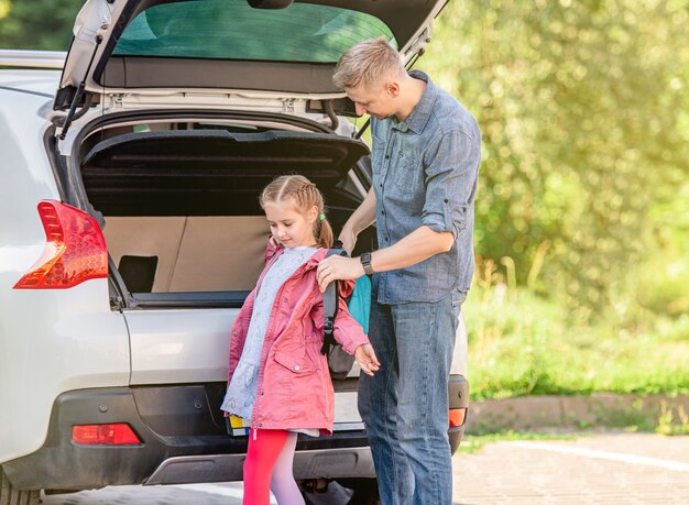 Pai ajudando a tirar a mochila da filha depois da escola perto da mala do carro