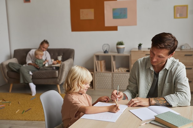 Pai ajudando o filho a fazer o dever de casa na mesa com a mãe brincando com o filho pequeno