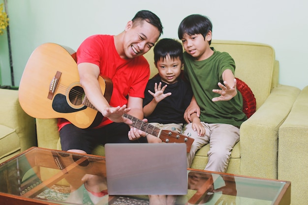 Pai asiático com guitarra sorrindo junto para o laptop com o filho durante a videoconferência