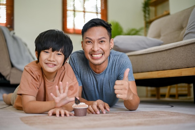 Pai asiático feliz mostrando o polegar para cima enquanto estava deitado no chão da sala com seu filho