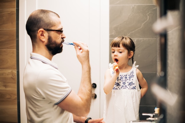 Pai barbudo e sua filha escovando os dentes em um banheiro. homem ensinando a filha a escovar os dentes.