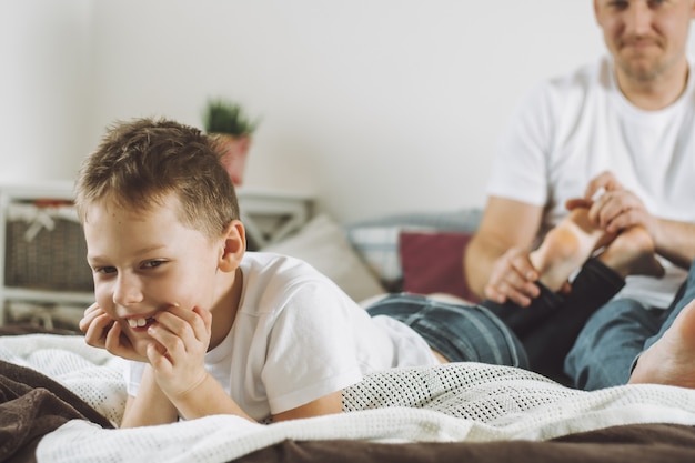 Pai brinca com seu filho 7-10 na cama. Papai faz cócegas nos pés das crianças. Família se divertindo