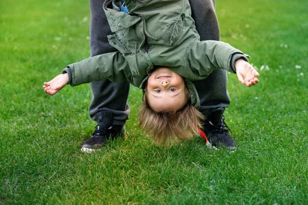 Pai brincando com o filho ao ar livre em uma grama. Menino da criança balançando de cabeça para baixo. Criança feliz em um parque. Conceito ativo de parentalidade.