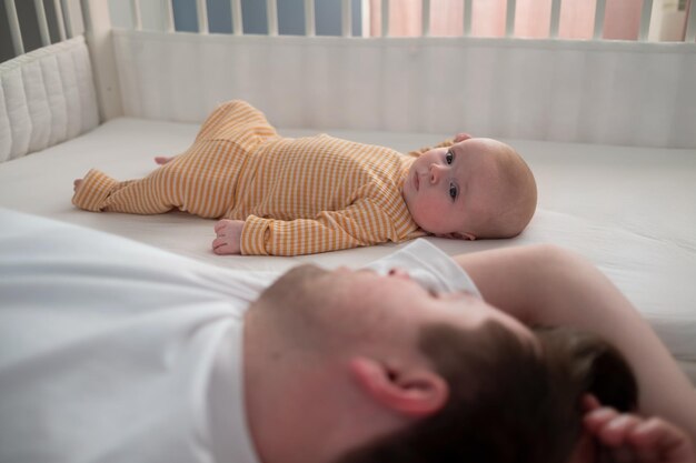 Foto pai cansado dormindo e seu filho recém-nascido deitado na cama olhando para a câmera em casa