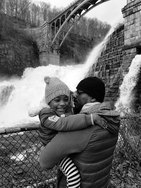 Foto pai carregando filha enquanto está de pé contra a cachoeira durante o inverno