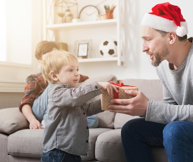 Pai com chapéu de Papai Noel dando ao filho pequeno um presente de Natal em casa na sala de estar