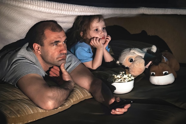 pai com filha assistindo a um filme no sofá à noite, pipoca na frente deles