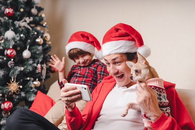 Pai com filho e cachorrinho com chapéu de Papai Noel em um chat por vídeo