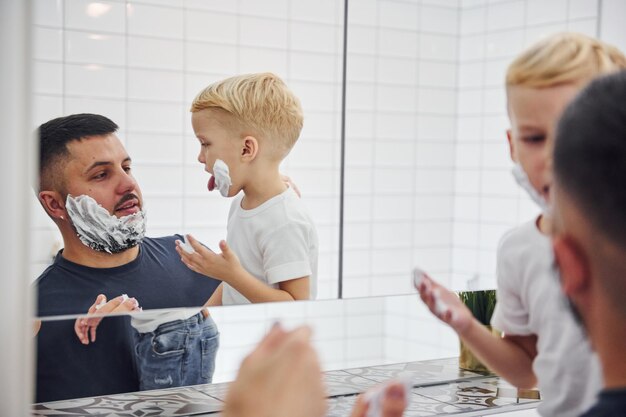 Pai com filho está no banheiro se divertindo usando gel de barbear e olhando no espelho.