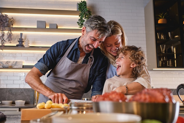 Pai de cabelos grisalhos no avental com filho pequeno encaracolado e esposa sorridente juntos e se divertindo enquanto preparava a refeição