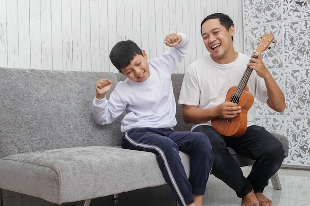 Pai de camiseta branca está tocando ukulele enquanto canta com seu filho de suéter está dançando