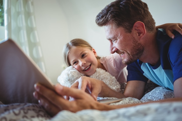 Pai deitado com a filha na cama e usando tablet digital