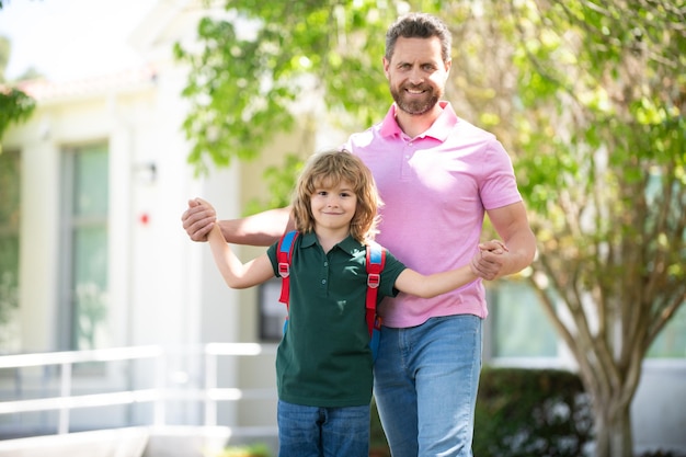 Pai e aluno da escola primária andam de mãos dadas Professor de camiseta e estudante bonito com mochila perto do parque escolar Pai leva um menino da escola infantil na primeira série