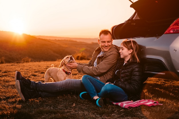 Pai e filha com cachorro sentado ao lado do carro na colina