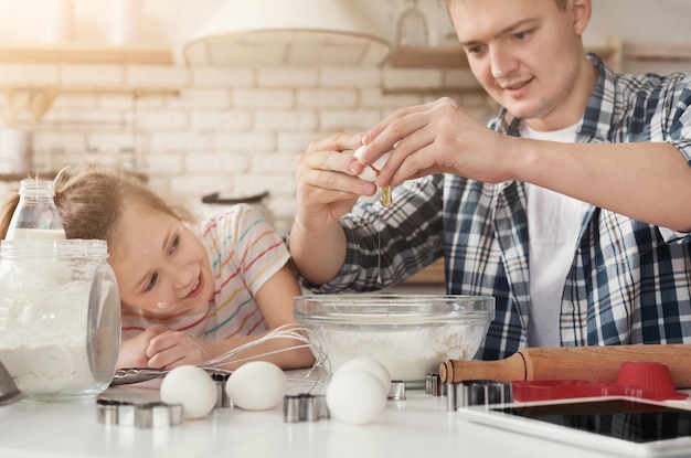 Pai e filha cozinhando bolo surpresa caseiro para a mãe. Menina olhando como seu pai quebrando ovo para massa em farinha, copie o espaço