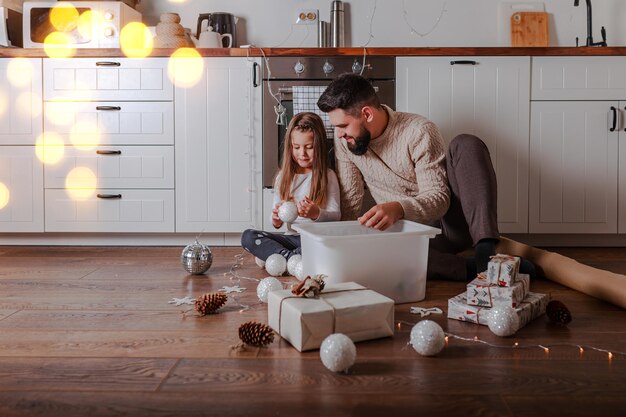 Pai e filha decoram os presentes de natal em casa. família abre brinquedos de natal no chão de madeira.