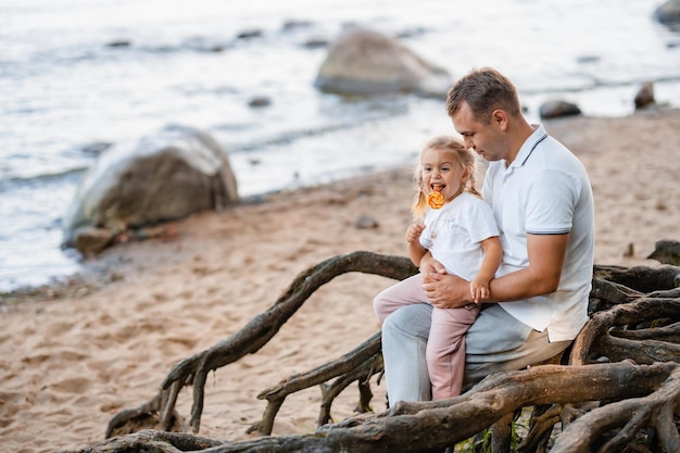 Pai e filha loira bonitinha juntos no verão na praia na natureza uma garotinha come raízes de uma árvore de pirulito